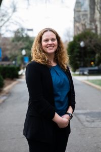 Dr. Jessica Ballou stands with her hands intertwined in front of her. She's standing on a walkway on Georgetown's main campus wearing a black blazer, blue shirt and black pants. Behind her is Healy Hall and a gray sky.