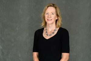 A professor stands against a gray background. She wears a black v-neck shirt and a necklace. Her arms are crossed in front of her. She has blonde hair and is smiling.