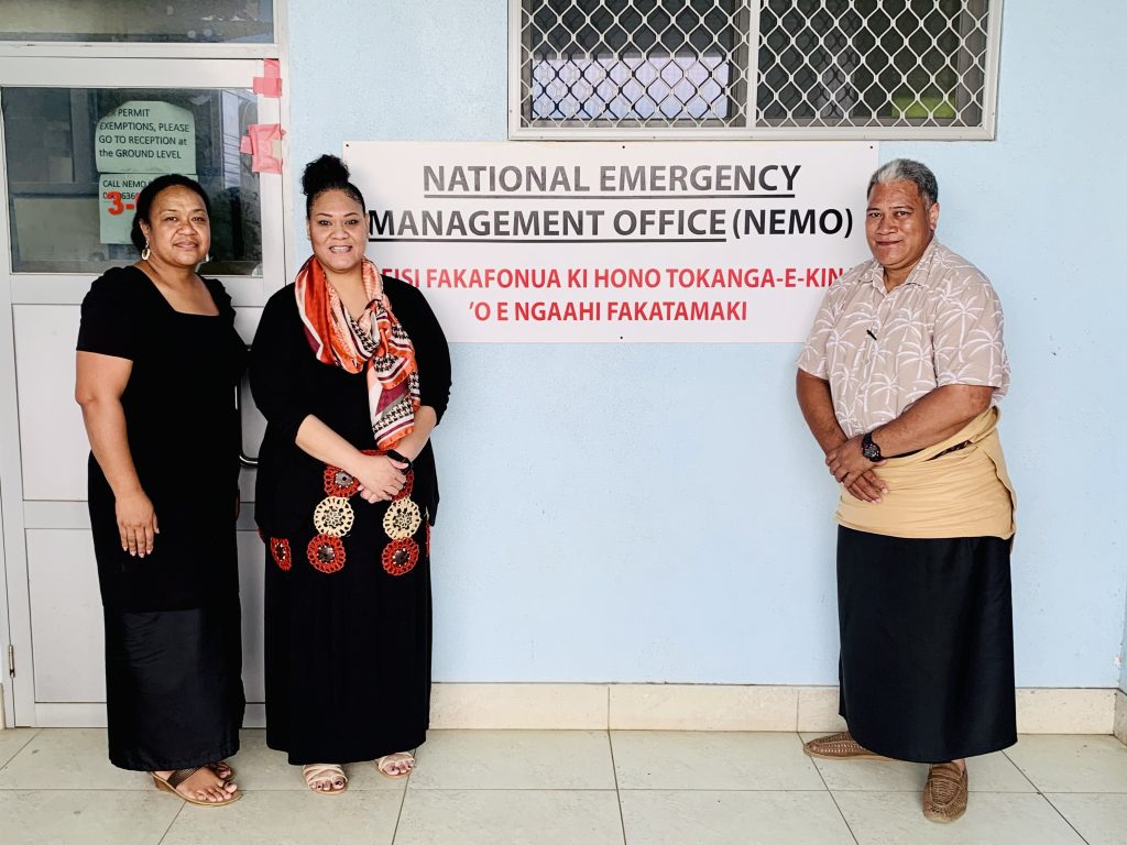 Lavinia Taumoepeau-Latu (G’23) (second from left) stands in front of a blue wall with a sign on it that says "National Emergency Management Office" in Tonga. She wears a black dress with a scarf, and stands next to the Principal Assistant Secretary Moana Fakava-Kioa (far left) and NEMO Director Mafua Maka (right).