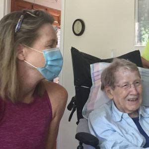 Carole Roan Gresenz sits next to her mother, who is in a wheelchair with two pillows propped next to her. Gresenz wears a pink sleeveless top and a blue mask. Her mother is smiling, wearing glasses and a blue blouse.
