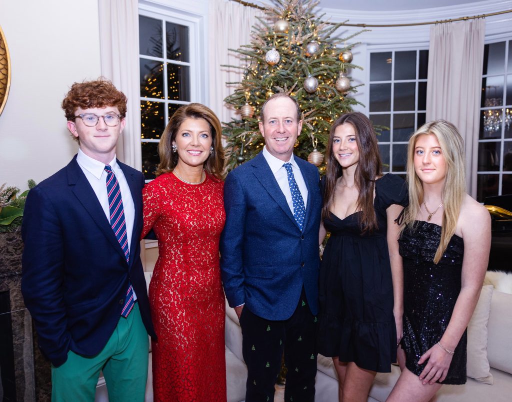 An image of Norah O'Donnell and Geoff Tracy (center) standing together with their three children in front of a Christmas tree inside a home. Norah wears a red dress, her son (left) wears a black blazer suit and green pants. Geoff wears a blue suit, and their two daughters (right) wear black dresses.