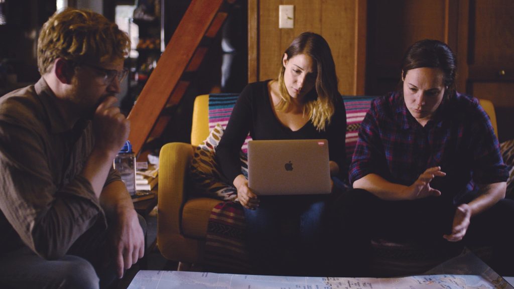 Still from film, three individuals sitting around table, one holding laptop computer.