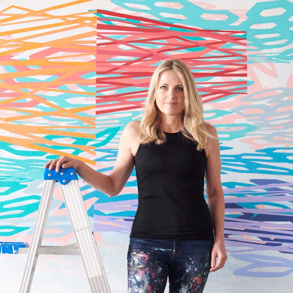 Rebecca Rutstein in black top and paint-covered jeans, standing next to ladder in front of painted backdrop.