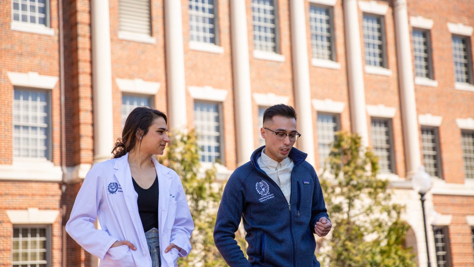 A female student in a white coat and a male student walk on a college campus