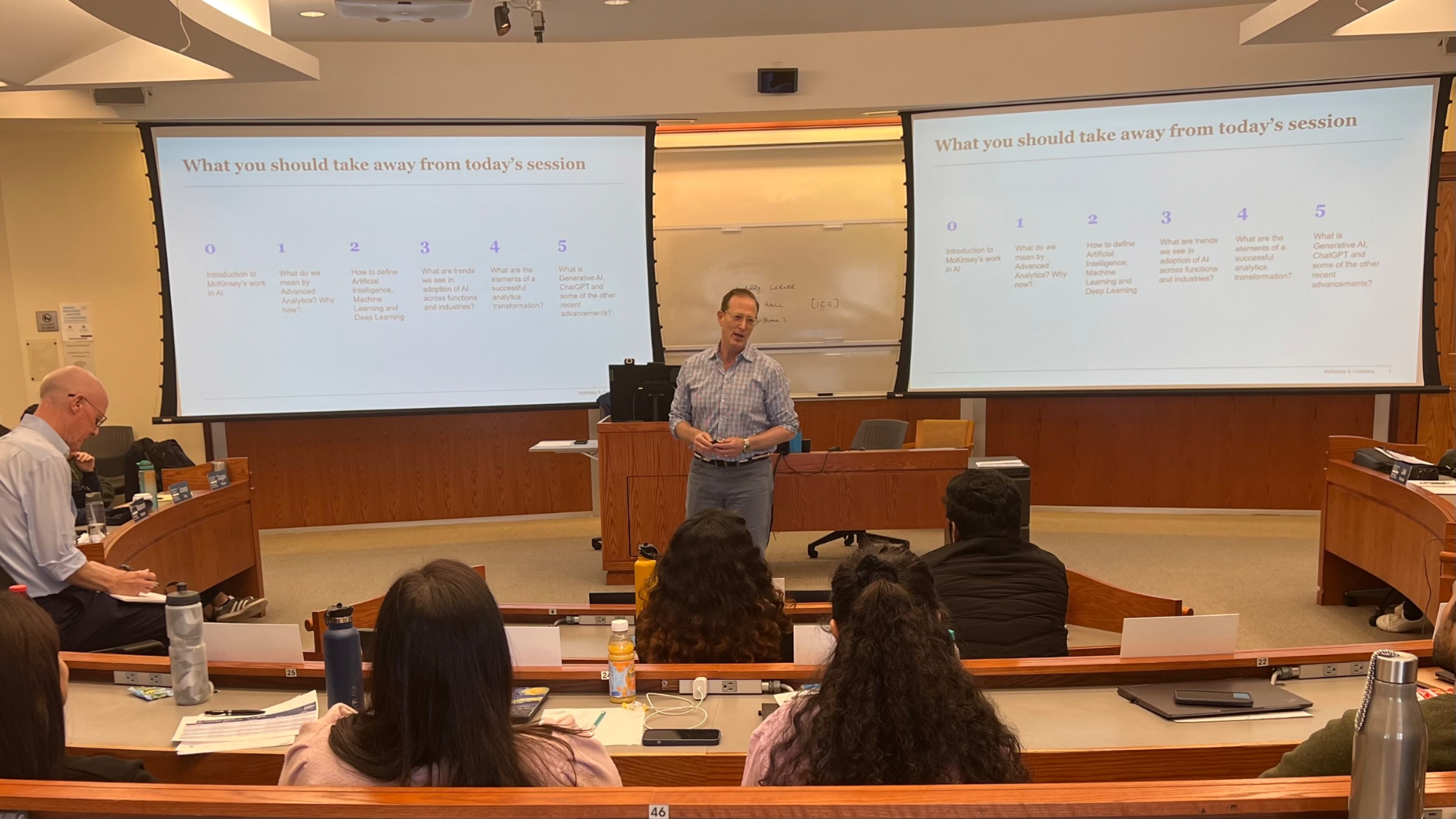 Person standing in front of lecture hall, with powerpoint in background.