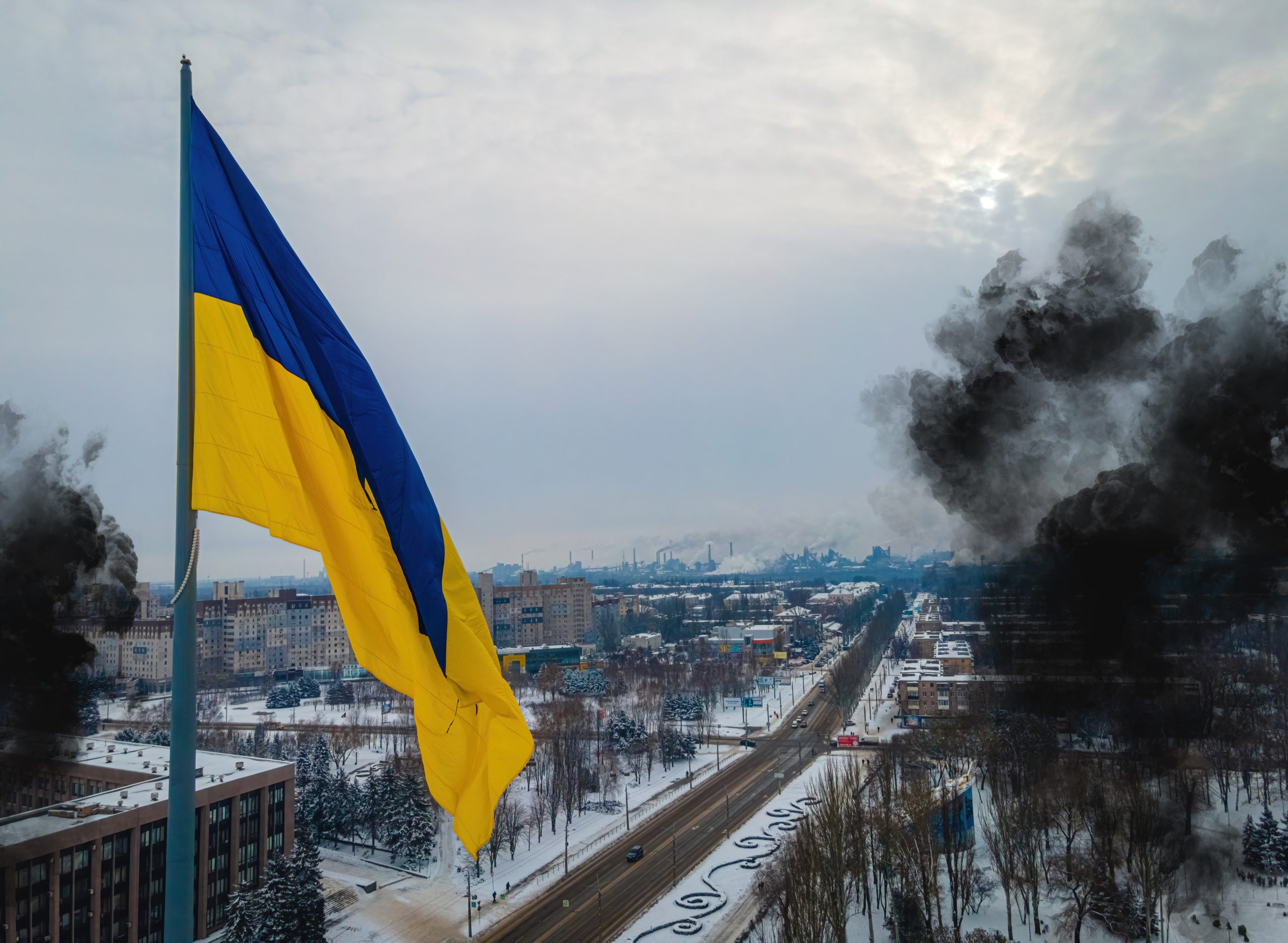 An image of the Ukrainian blue-and-yellow flag hanging over a city in Ukraine. Behind it is black smoke from an explosion and a cloudy sky.
