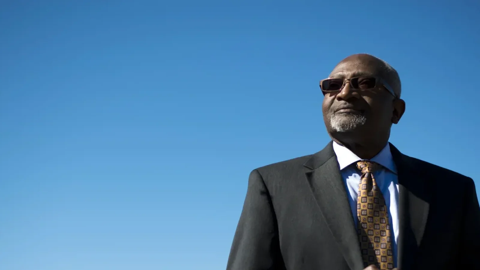 Bllard in suit with sunglasses, against blue sky.