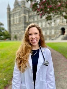 Lauren Havens is a medical student at Georgetown. In this picture, she wears a white coat and a stethoscope and stands in front of Healy Hall and its green lawn. 