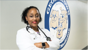 Michelle Roett, founding director of the Center for Health Equity, folds her arms and stands in front of a seal that says Georgetown Medicine. She wears a white coat and a stethoscope.