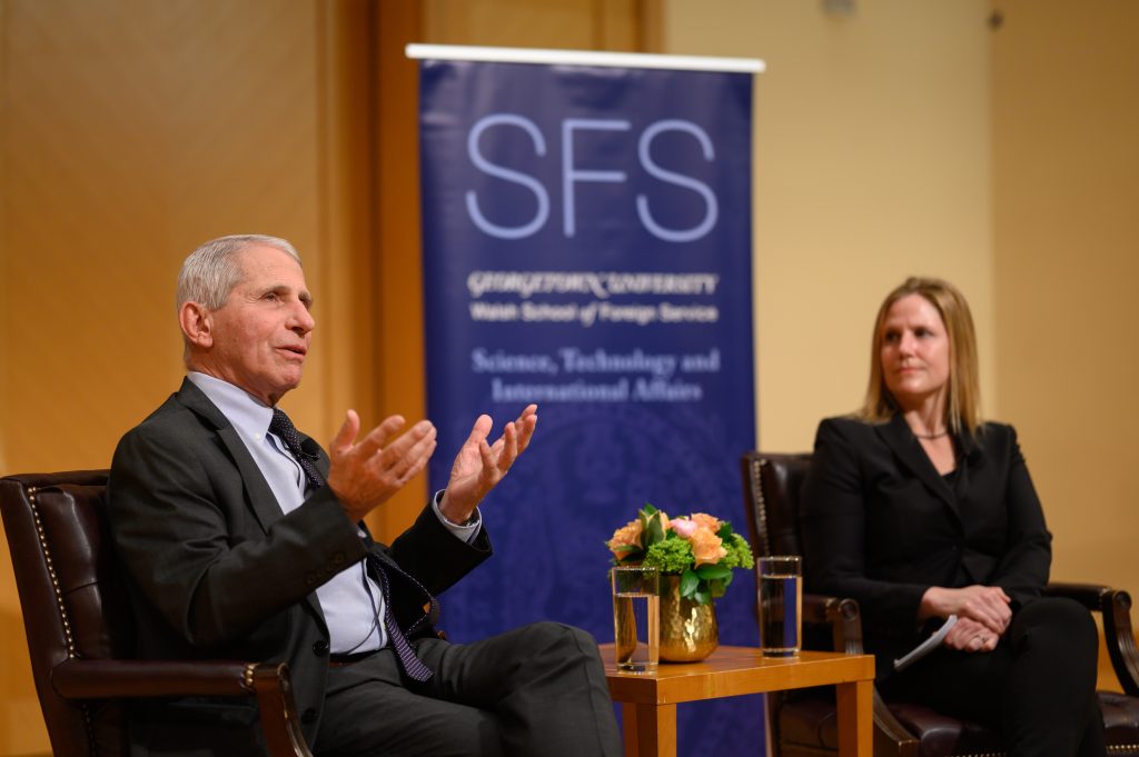 Dr. Fauci sits on stage in a panel discussion. He wears a suit and gestures with his hands as he speaks. Next to him are the panel moderator and a small table with a bouquet on it. 