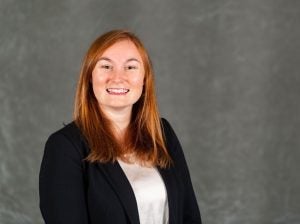 A headshot of Brittany Fried, who has red hair and is wearing a black suit with a white top. She is smiling at the camera.