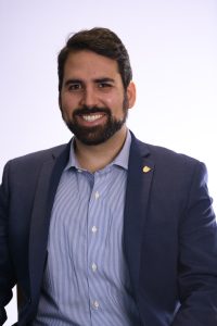 A headshot of Carlos Chacon, a Presidential Management Fellow. Carlos wears a navy suit with a blue and white collared shirt underneath. 