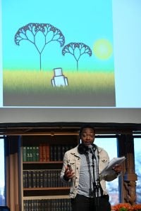 A student performs at Georgetown's ACDC: Arts Celebrating Disability Culture event. He stands on stage in Riggs Library and speaks into a microphone. Behind him is the animation he created.