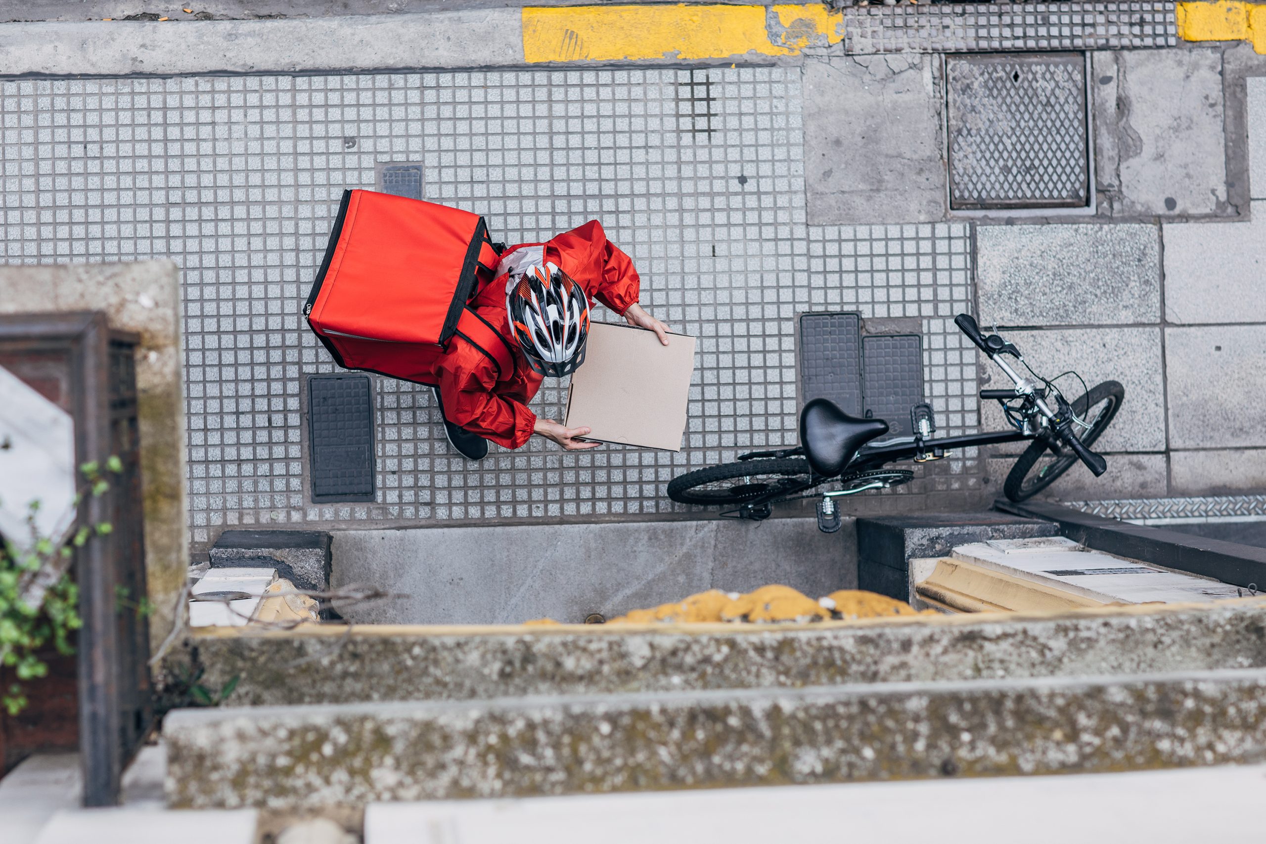 A view looking down from a building on a food delivery driver, who wears a red jacket and carries a red delivery pack on his back. Next to him at the entrance to a building is his bicycle.