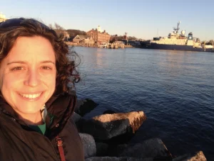Rebecca Helm pictured with a harbor in the background with a boat and some buildings.