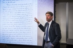 Professor Adam Rothman, founding director of the Center for the Study of Slavery and Its Legacies, stands in front of a screen with a document on it. He holds up one hand and talks, wearing a suit jacket and tie.