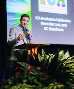 Jelenick speaks at Western Carolina University in December 2019 at the Intercultural Affairs (ICA) Fall Graduation Celebration. He stands behind a podium holding a microphone and wears a gray suit. Behind him is a screen.