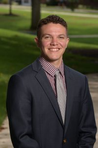 A headshot of Riley Jelenick, the new director of Georgetown's LGBTQ Resource Center. Riley wears a suit with a button-up shirt and gray tie. He smiles at the camera in front of a green lawn.