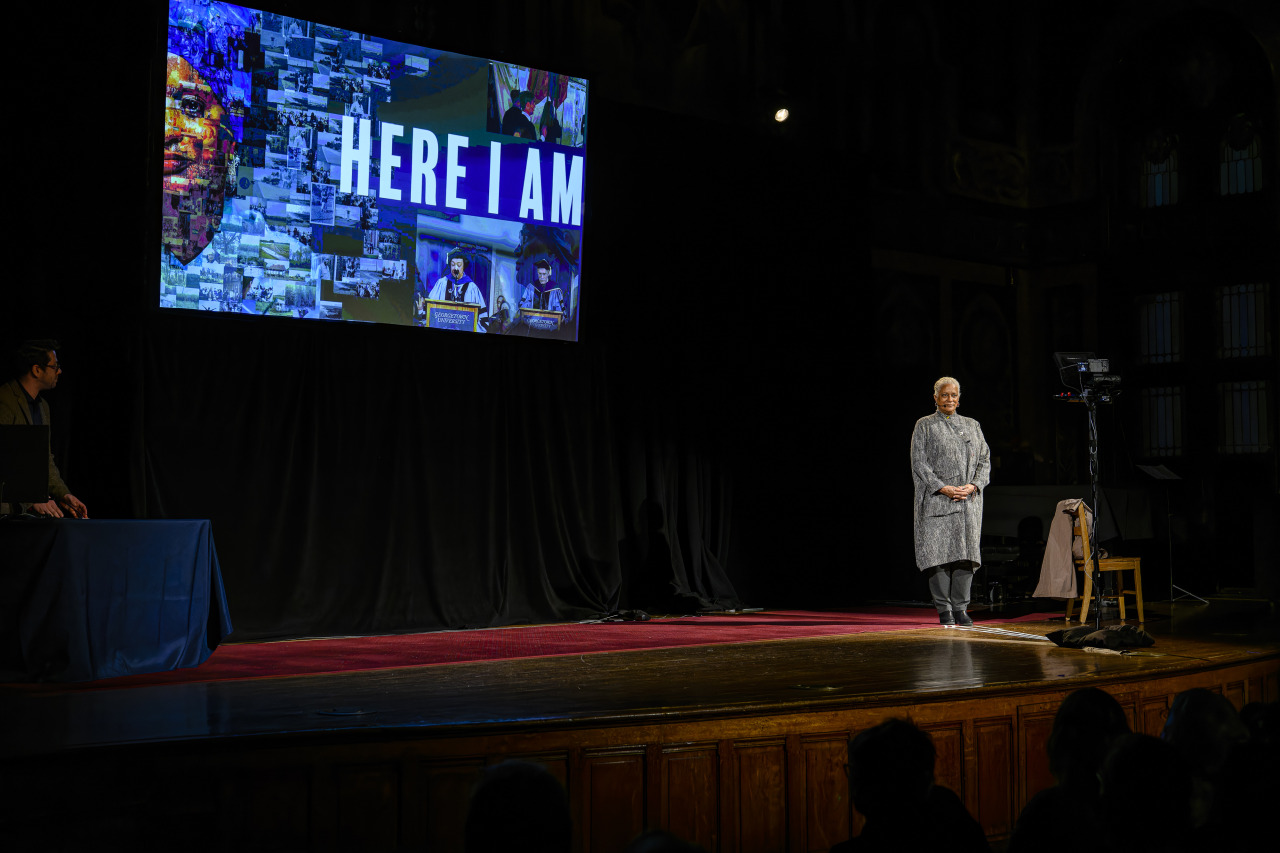 Mélisande Short-Colomb stands on a stage with her hands crossed in front of her. Behind her is an illuminated screen that says "Here I am," the title of her performance.