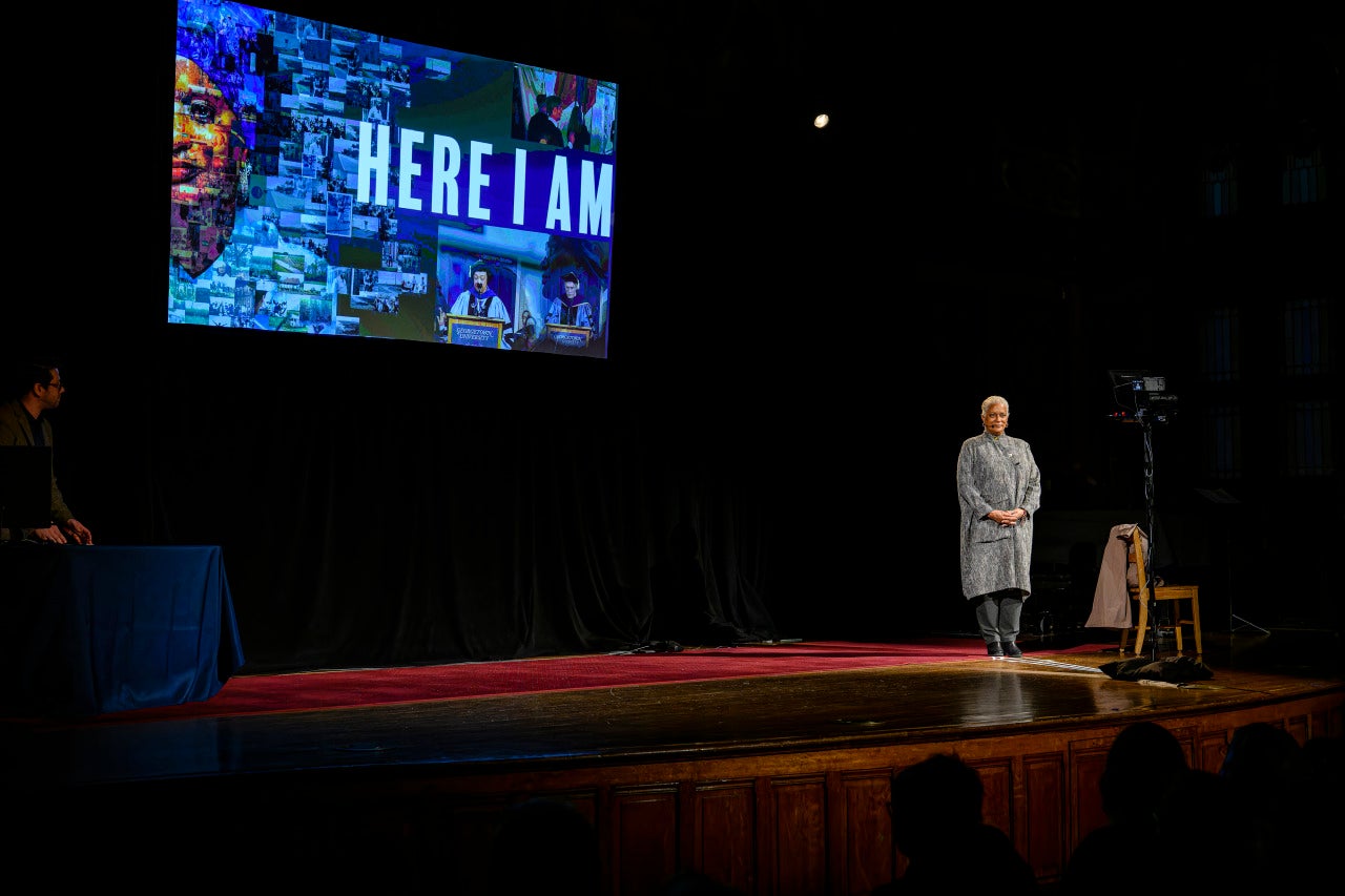 Mélisande Short-Colomb stands on a stage with her hands crossed in front of her. Behind her is an illuminated screen that says "Here I am," the title of her performance.