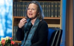 Carolyn Forché, a Distinguished University Professor in the Department of English, sits on a chair with books behind her. She is wearing glasses and a blue scarf and holds up one arm as she speaks.