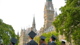 A group of graduating seniors wearing their cap and gowns look at Healy Hall, with its clocktower in the background.