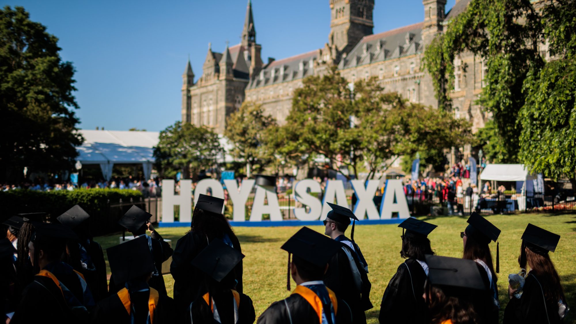 A sign that says &quot;Hoya Saxa&quot; sits on Healy Lawn in the background. Students in regalia are in the foreground.