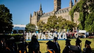 A sign that says &quot;Hoya Saxa&quot; sits on Healy Lawn in the background. Students in regalia are in the foreground.