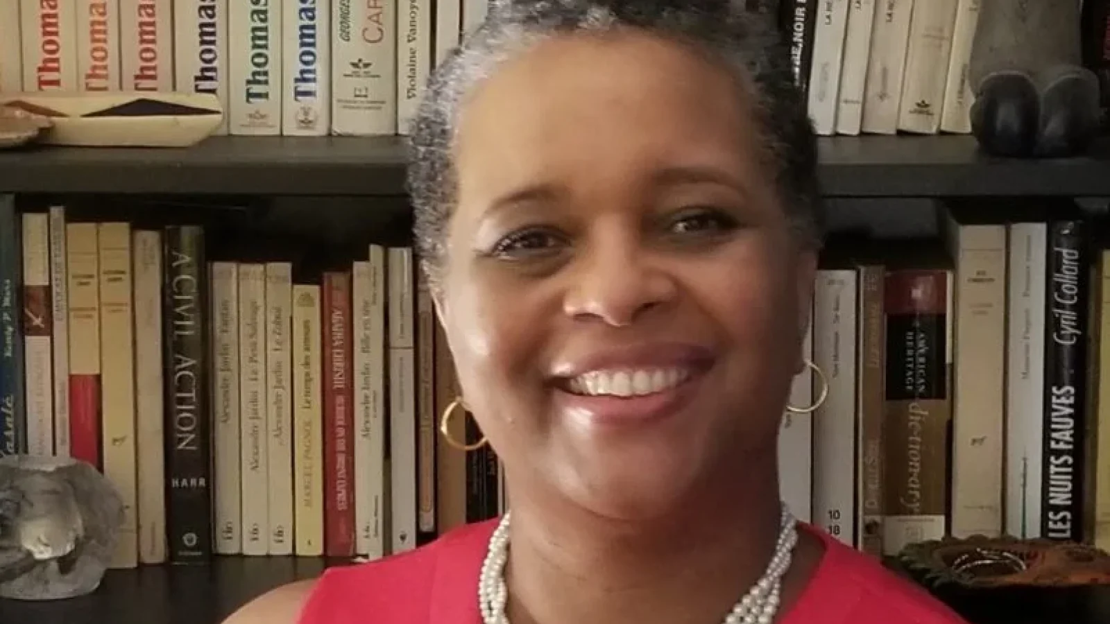 Carline Noailles (EMBA&#039;23) smiles at the camera. She wears a bright pink sleeveless shirt and pearls and stands in front of a bookshelf.
