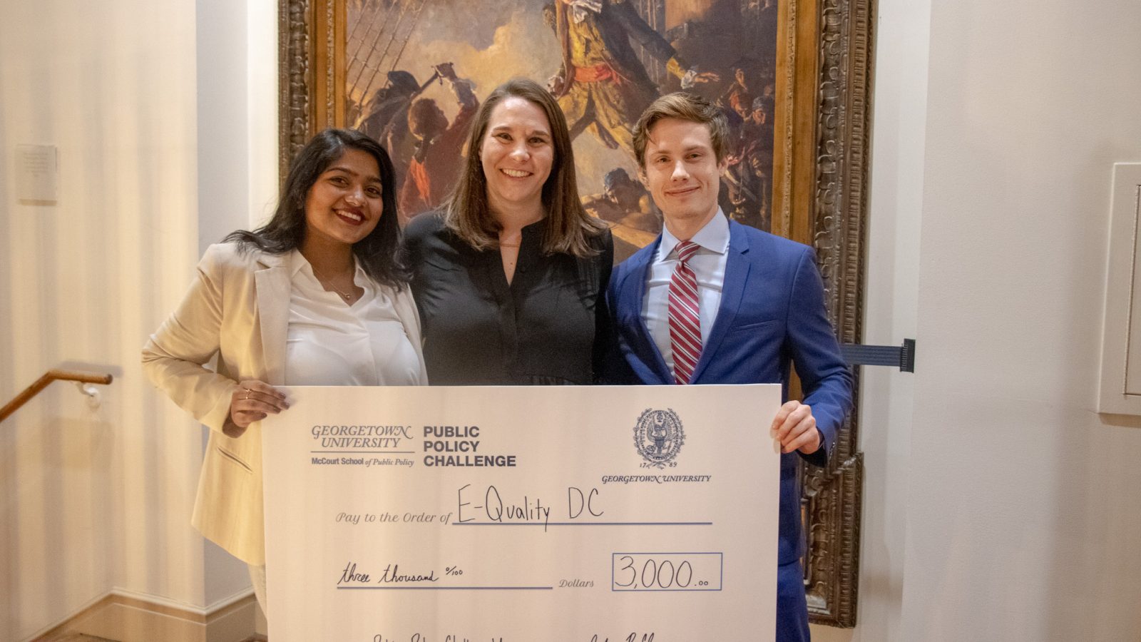 Three winners of the Georgetown Policy Challenge stand side-by-side holding a check. Behind them is an oil painting.