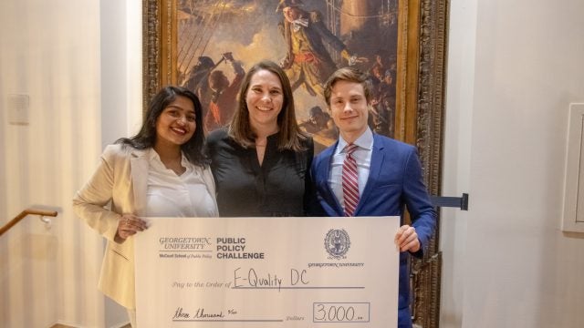 Three winners of the Georgetown Policy Challenge stand side-by-side holding a check. Behind them is an oil painting.