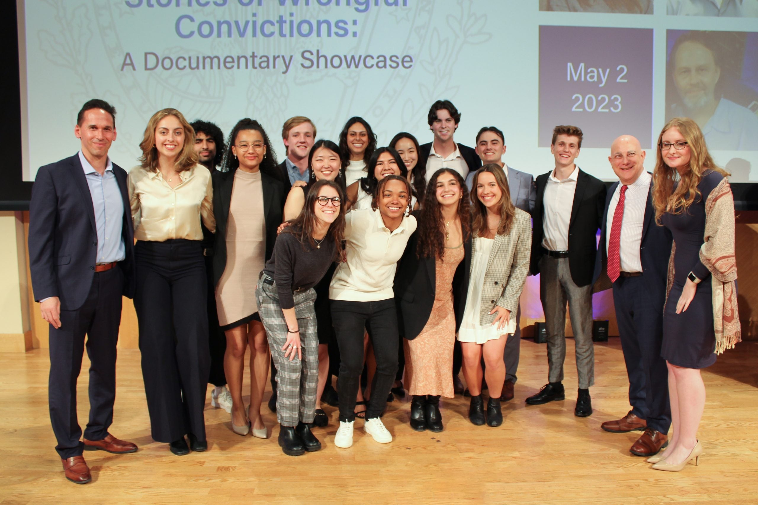 A large group of people including Cassera stands on stage with a projector screen behind them.