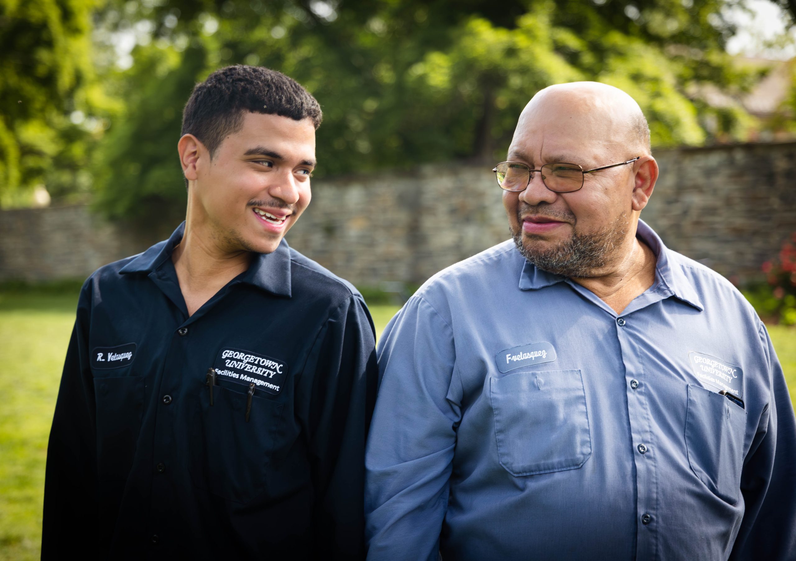 Reynaldo Velasquez stands with his back to his father, Francisco Velasquez, as they smile at one another. They both wear suits as part of Georgetown's facilities team and stand on the lawn outside.