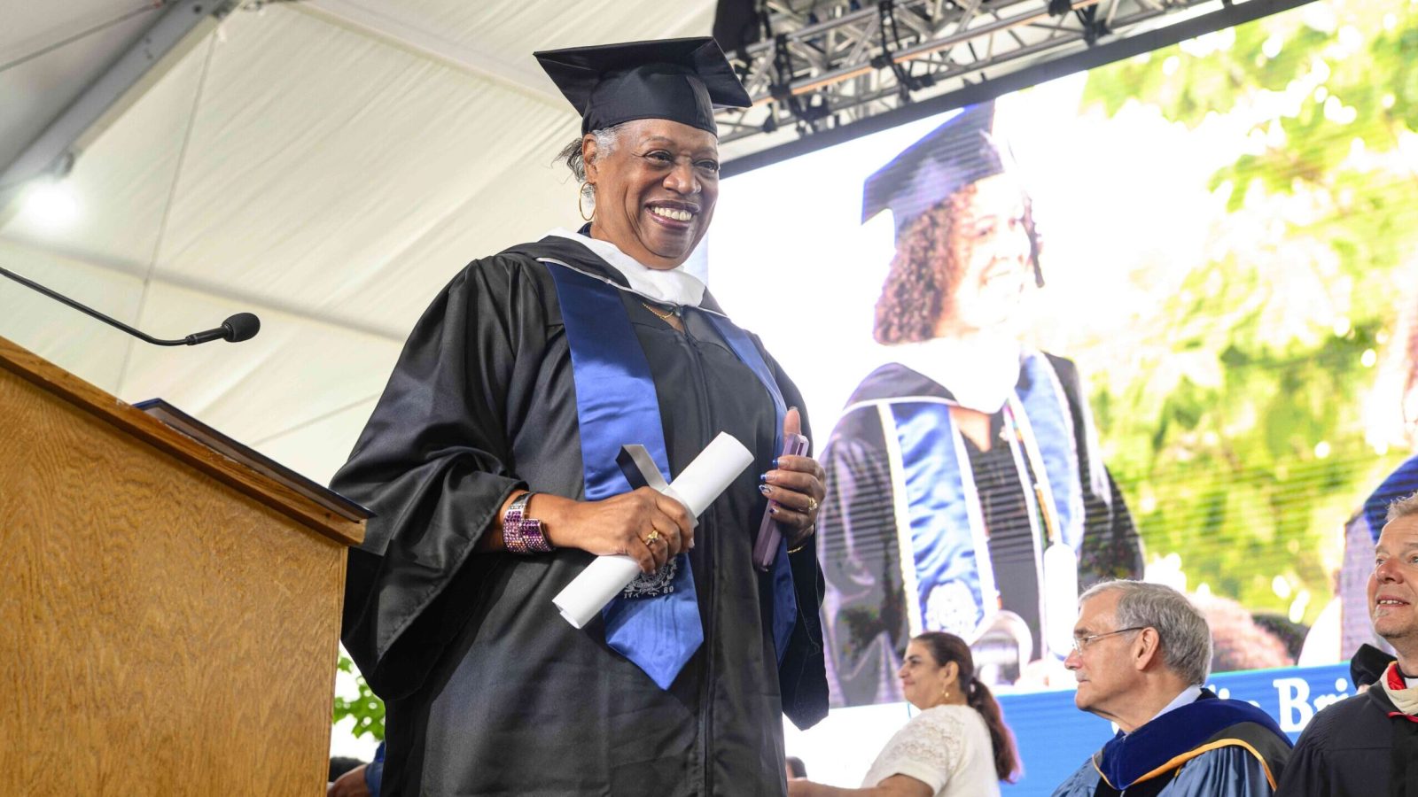 Karen Franklin walks across the stage with her degree in hand. She wears a cap and gown and smiles at the camera.