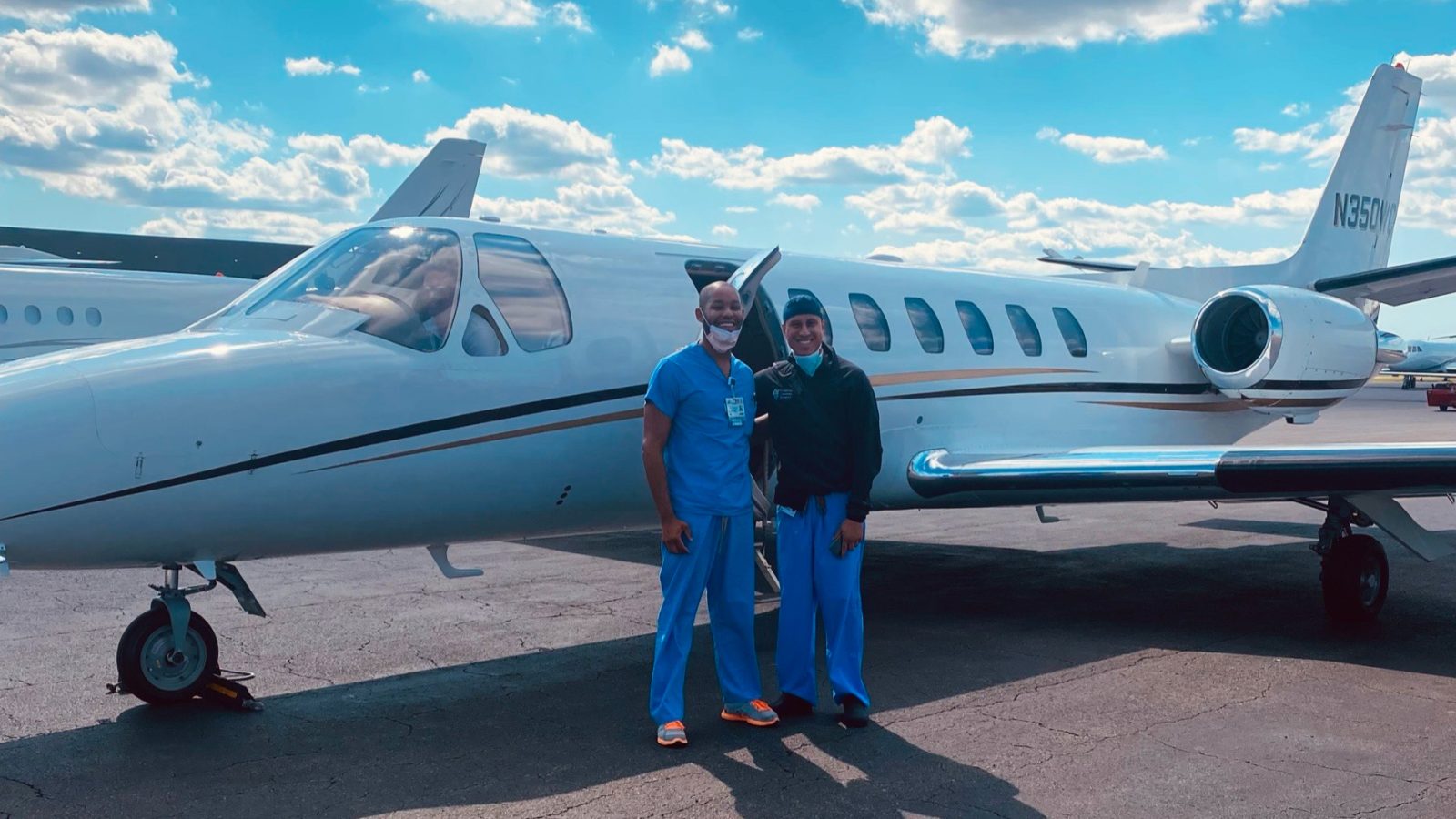 Malcolm standing with someone in front of a small plane while wearing doctor scrubs.