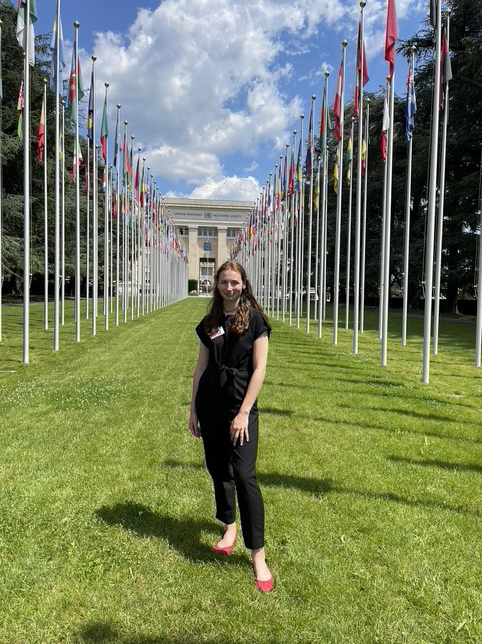 Hardy stands on a grassy lawn with flagpoles lining the lawn leading up to the UN building in Geneva.