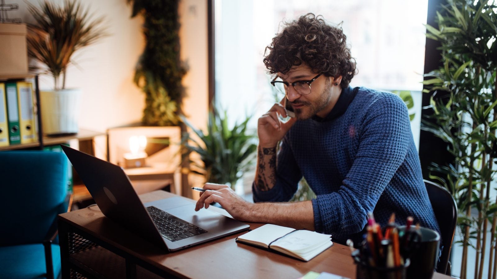 An employee talks on the phone while typing on his laptop at his home desk. Behind him are green plants.