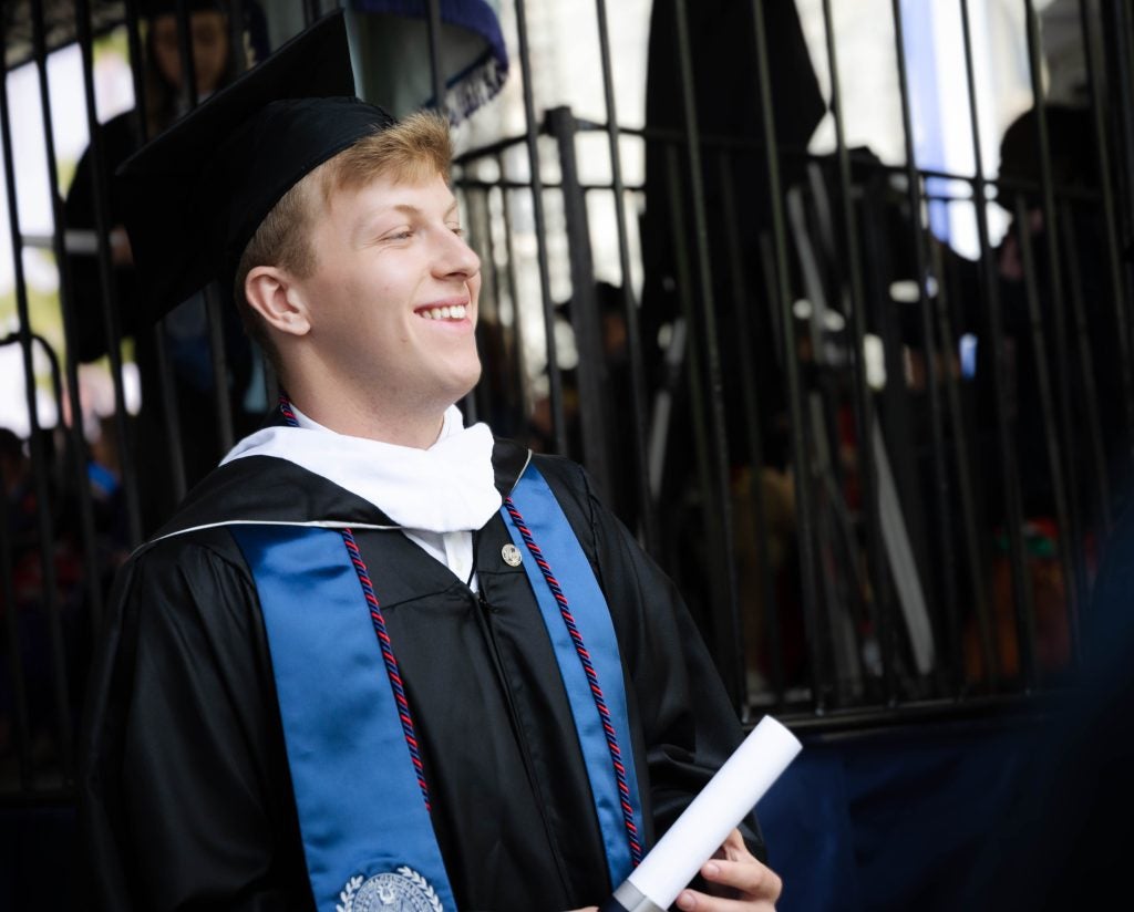 Jackson Mittleman (C'23) smiles on his graduation day. He's wearing a cap and gown and holding his diploma.