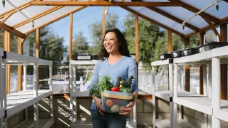 Naomi Hansen (B&#039;95) carries a basket of produce and stands outside in her greenhouse.
