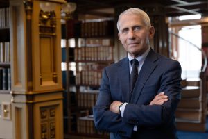Dr. Anthony S. Fauci stands with his arms crossed in Riggs Library on Georgetown's Main Campus. He wears a navy suit with a white shirt underneath and tie, and stands in front of bookcases.