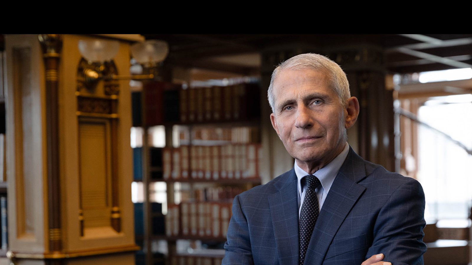 Dr. Anthony S. Fauci stands with his arms crossed in Riggs Library on Georgetown&#039;s Main Campus. He wears a navy suit with a white shirt underneath and tie, and stands in front of bookcases.