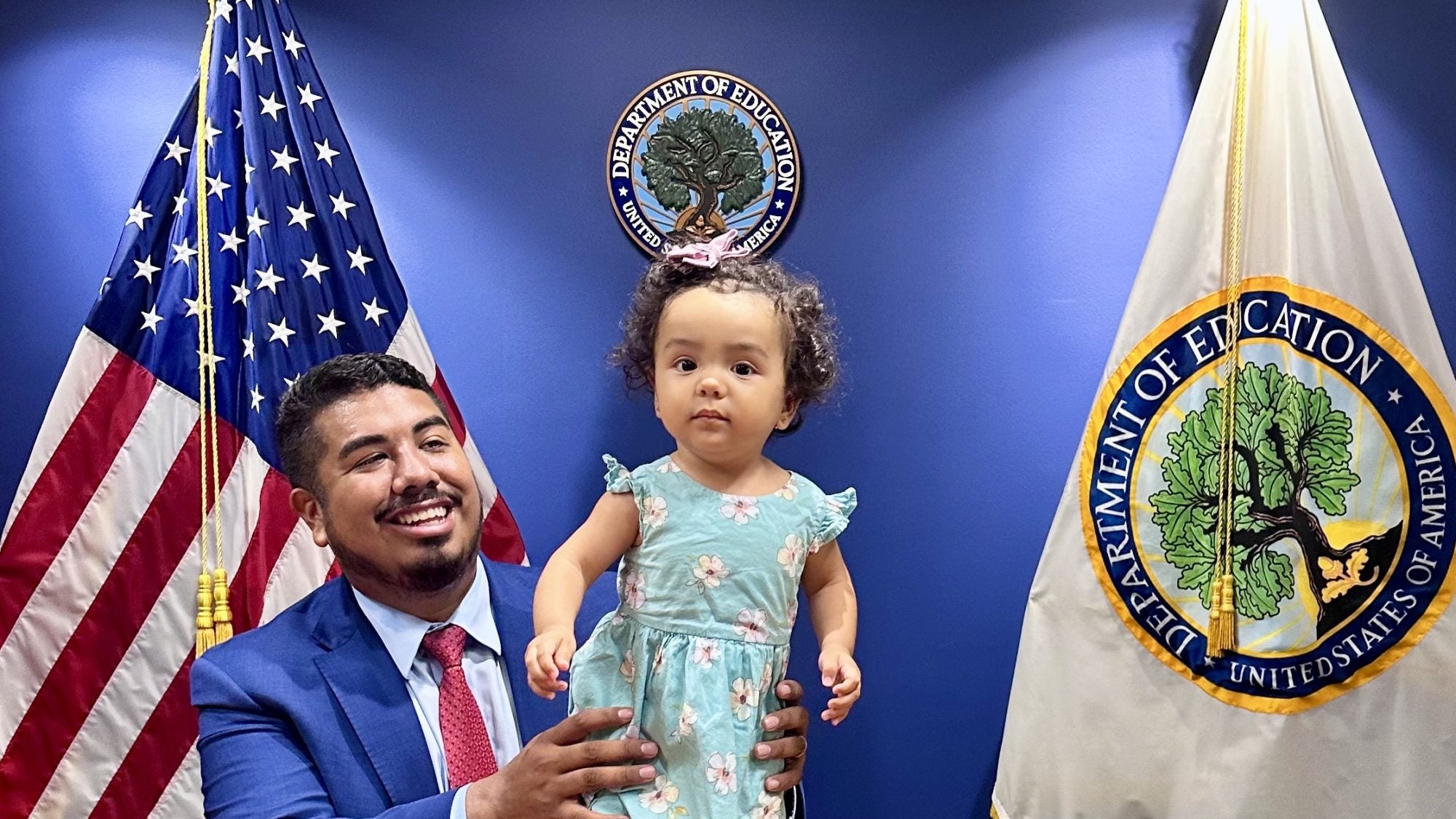 Adan Gonzalez (C&#039;15) holds up his daughter on the podium of a conference room at the U.S. Dept. of Education.