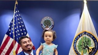 Adan Gonzalez (C&#039;15) holds up his daughter on the podium of a conference room at the U.S. Dept. of Education.