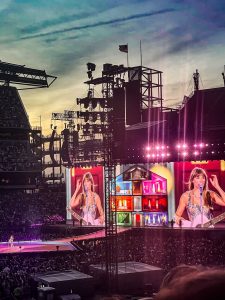 Taylor Swift on massive screens on a stage inside a stadium in the evening.