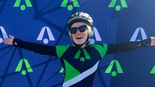 Marissa Nissley (B&#039;24) stands at the finish line after a 50-mile race with her arms outstretched. She wears black sunglasses and a helmet.