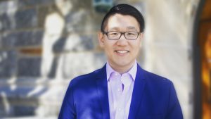 A headshot of Shawn Rhoads (G’22), a postdoctoral research fellow at the Icahn School of Medicine. Shawn smiles and wears a navy jacket, a purple collared shirt and glasses.