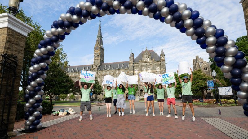 A group of Hoyas jump in the air beneath an archway of balloons and hold signs welcoming new Hoyas to the Hilltop campus.