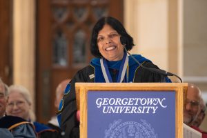 Reena Aggarwal, Robert E. McDonough Professor of Finance, speaks students at convocation. She smiles, wearing black academic robes, behind a podium. 