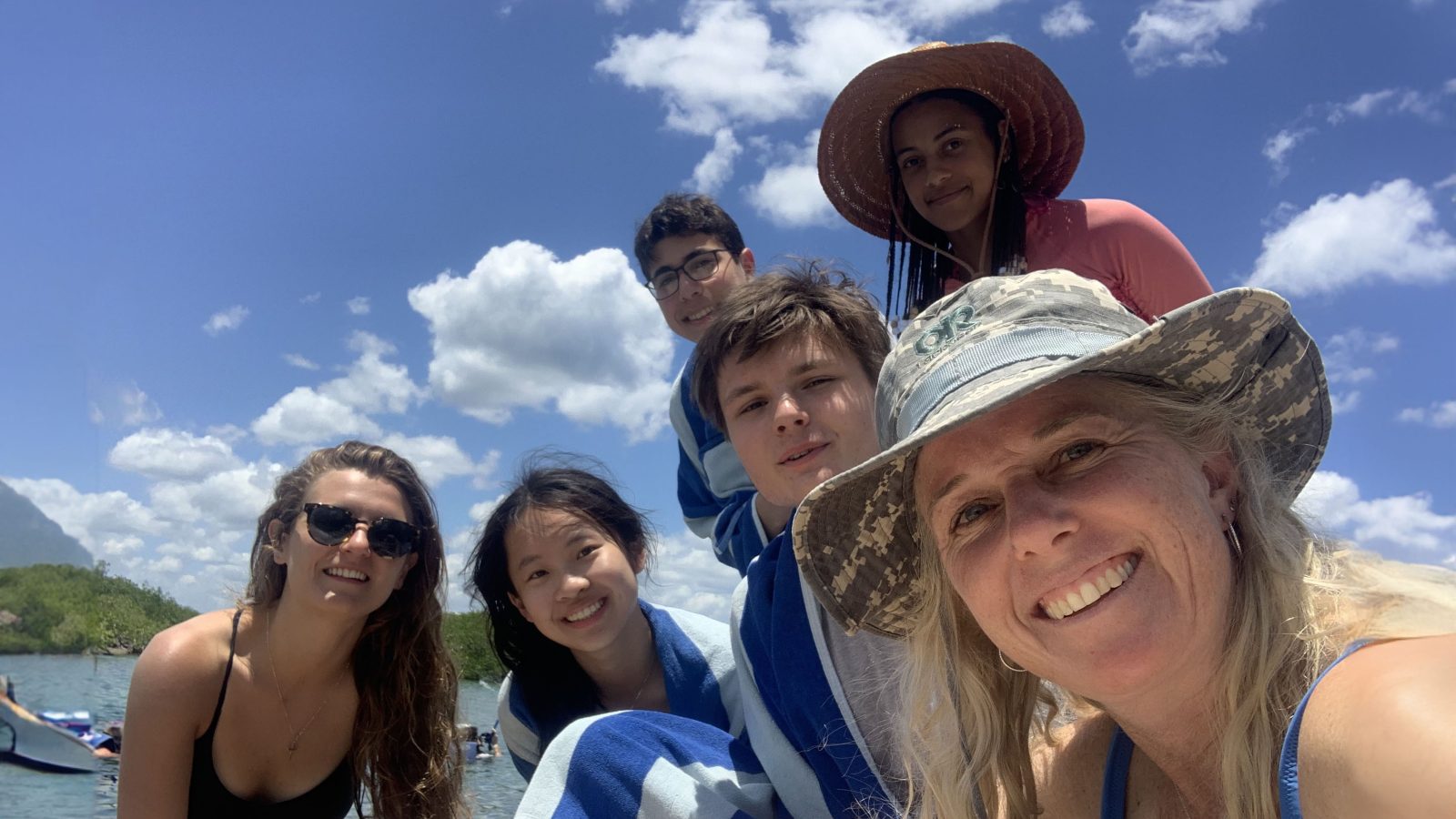 A group of students and a faculty member pose for a selfie with a beach and blue sky in the background.