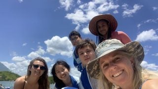 A group of students and a faculty member pose for a selfie with a beach and blue sky in the background.